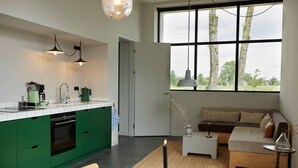 Una cocina moderna con armarios verdes, una mesa de comedor con una vela y una zona de estar con sofá y ventana con vistas a árboles.