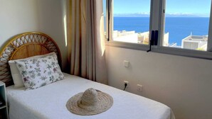 A bedroom with a bed, a window with a view of the sea, and a straw hat on the bed. - Villa Fénix Premium (Santa Cruz de Tenerife)
