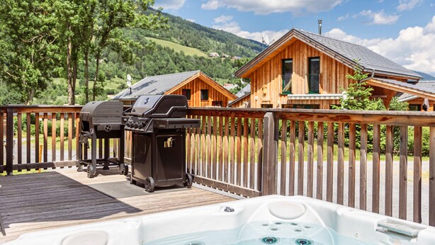 Une terrasse en bois avec un jacuzzi et des barbecues, entourée d’une clôture en bois et avec des maisons en arrière-plan.