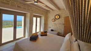 Une chambre avec un grand lit, un ventilateur de plafond, des murs en pierre et une vue sur la mer.