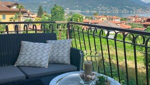 Ein Balkon mit einer Rattan-Couch, gemusterten Kissen, einem Glastisch mit Kerze und Muscheln sowie einem herrlichen Blick auf einen See und Berge.