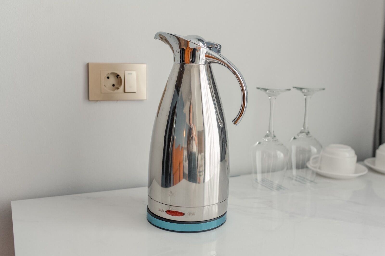 A stainless steel electric kettle on a white countertop with a power outlet and switch in the background.
