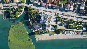 Vista aerea di una zona costiera con un porto turistico, un edificio con tetto circolare e edifici residenziali circostanti.