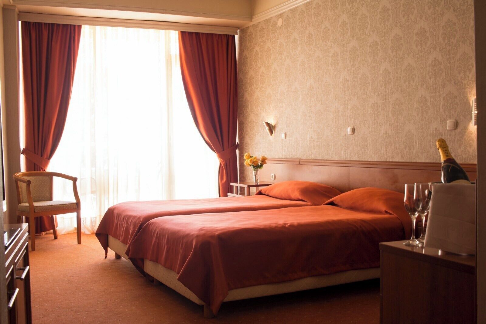 Habitació d'hotel amb llit de matrimoni, dos almoines, una cadira, una cambrera amb una ampolla de xampany i un ram de flors.