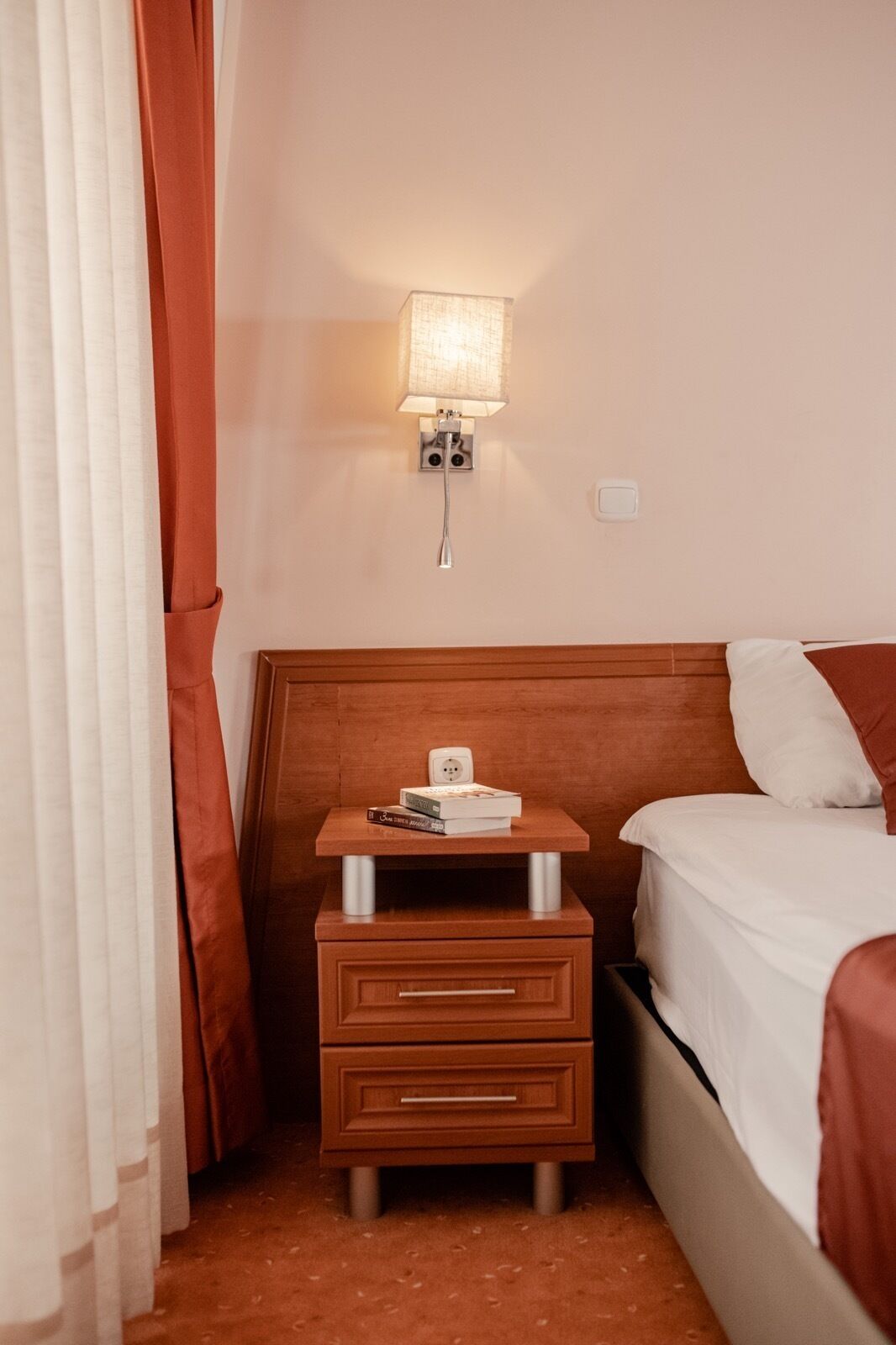 Une chambre d’hôtel avec une table de chevet en bois, une lampe, un lit avec une literie blanche et un rideau rouge.