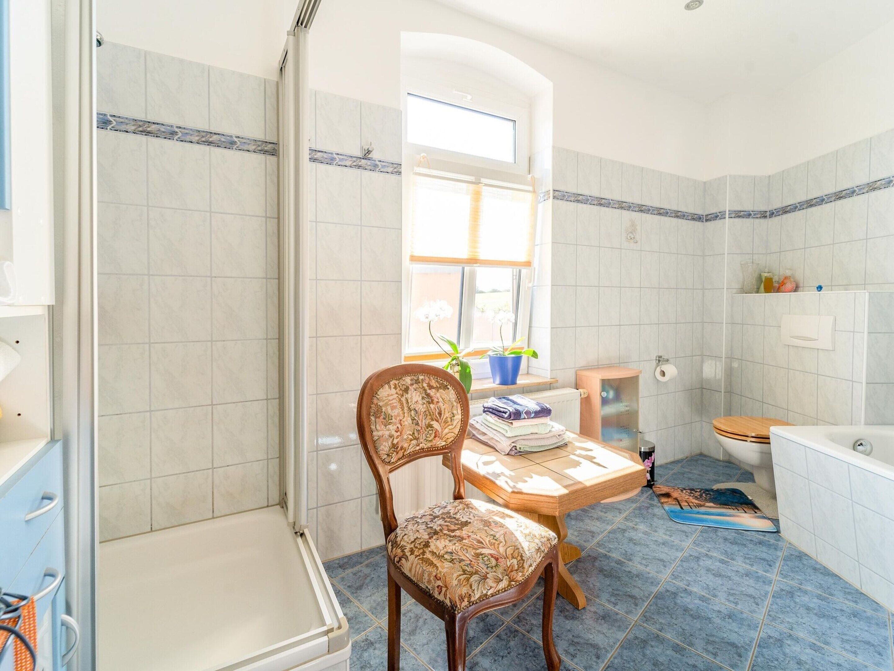 A bathroom with a shower, toilet, and a wooden chair with a floral cushion.