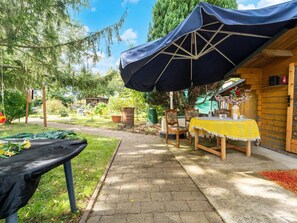 A patio area with a table and chairs under a large umbrella, surrounded by greenery and a wooden building. - Cozy Family Stay in Petersberg (Petersberg)