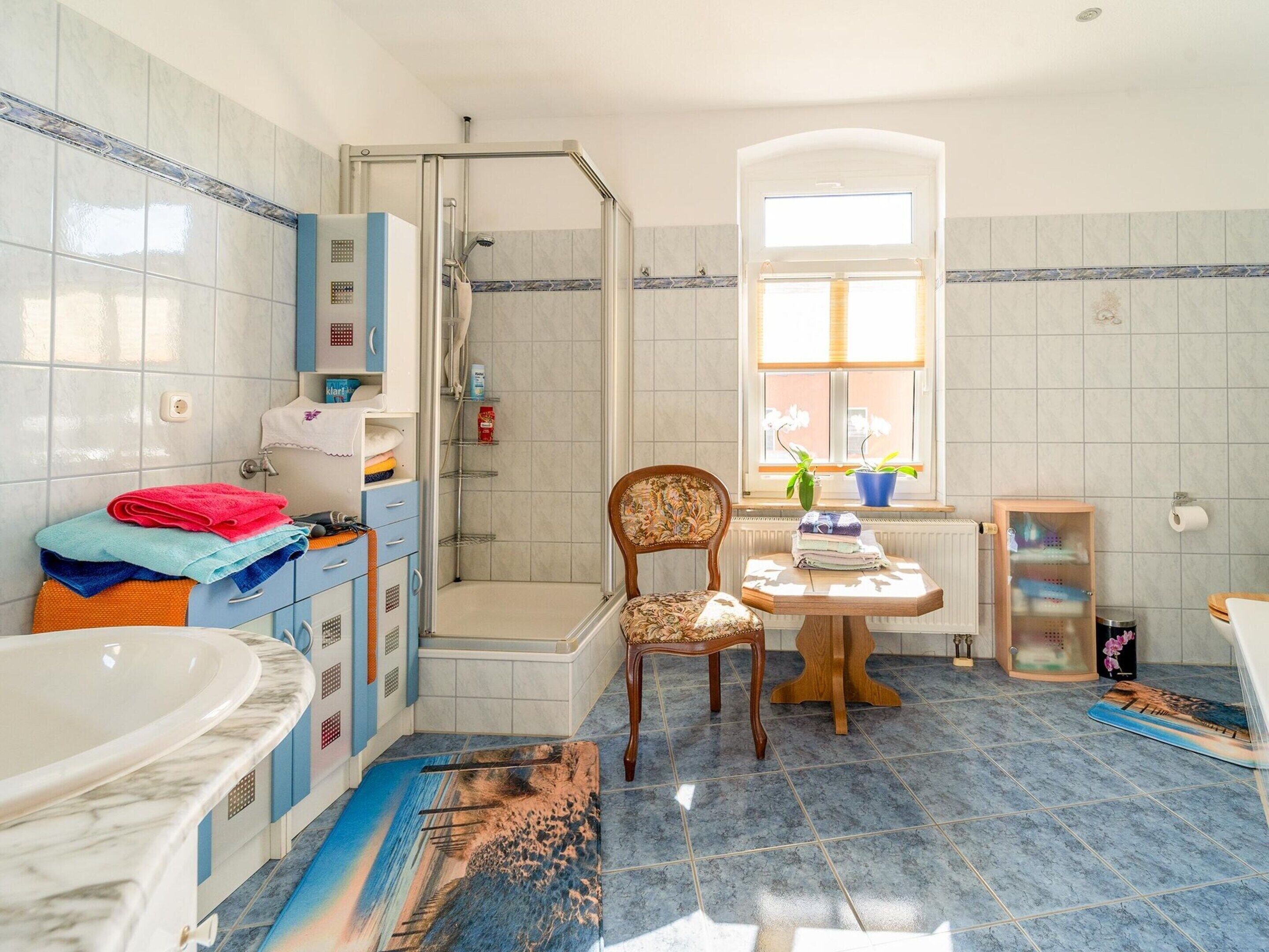 A bathroom with a shower, a sink, a chair, and a small table.