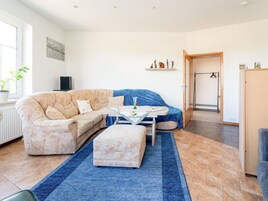 Uma sala de estar acolhedora com um sofá bege, uma poltrona azul, uma mesinha com toalha florida e um tapete azul.
