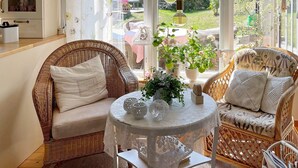 Una habitación acogedora con muebles de mimbre, una mesa redonda con objetos decorativos y vistas al jardín a través de ventanas amplias.