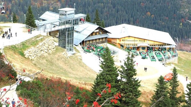 Een berglodge met een glazen lift, terras en een skilift op de achtergrond.