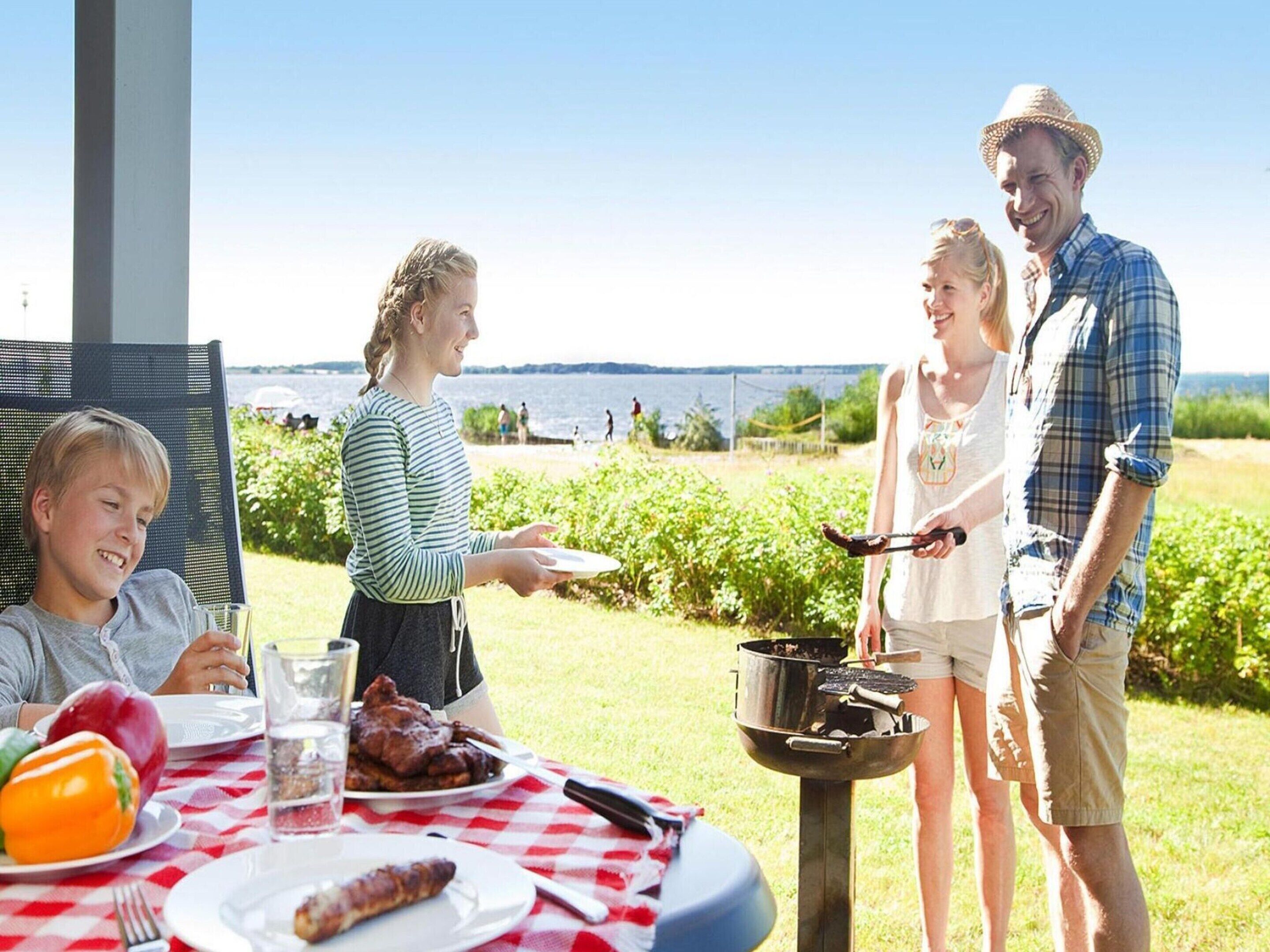 Un groupe de personnes est assis à une table en plein air, dans un espace aménagé pour les repas, avec vue sur l’eau et la verdure.
