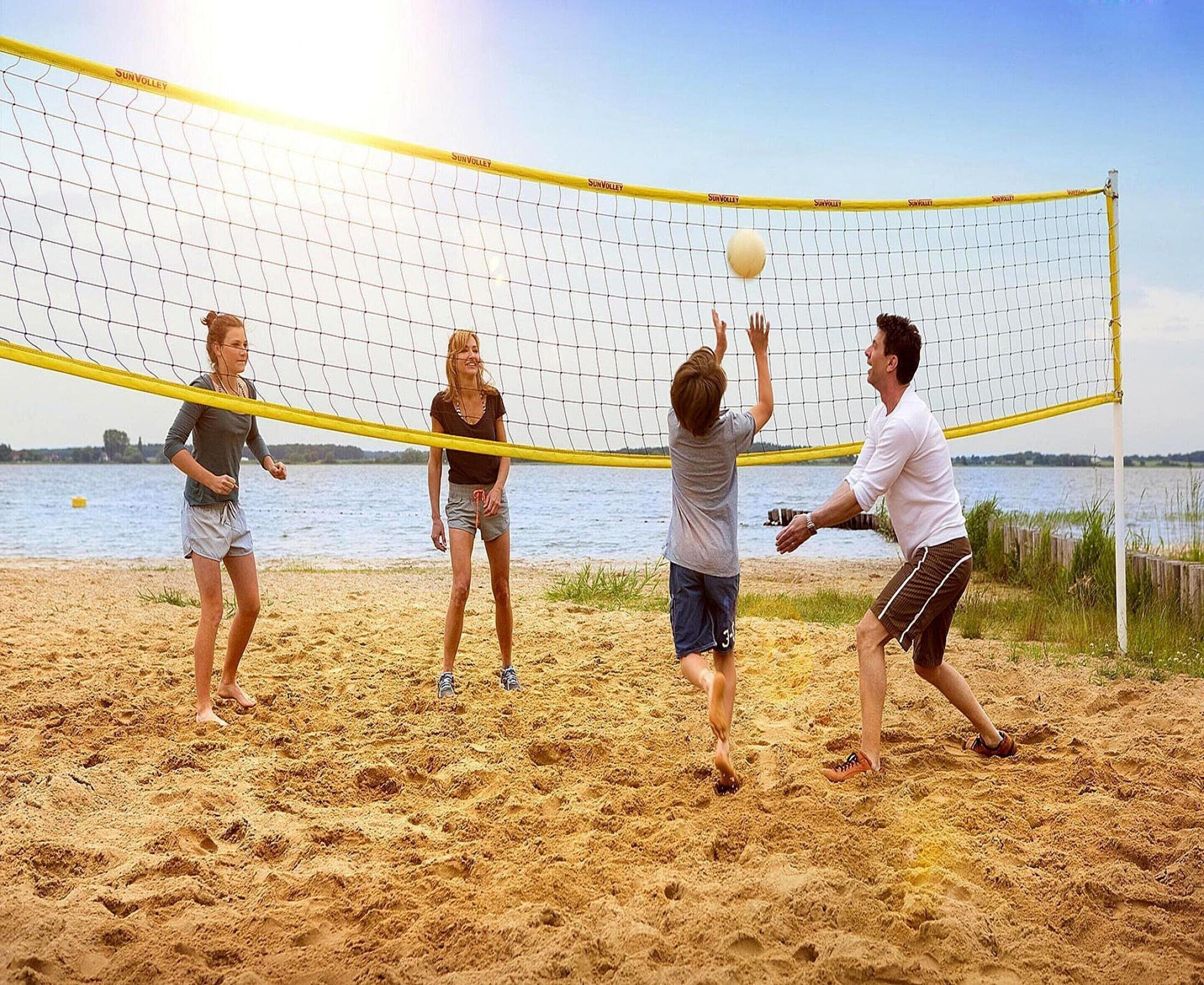 En grupp människor spelar strandvolleyboll på en sandplan med ett nät, ett vattenområde och en klar himmel i bakgrunden.