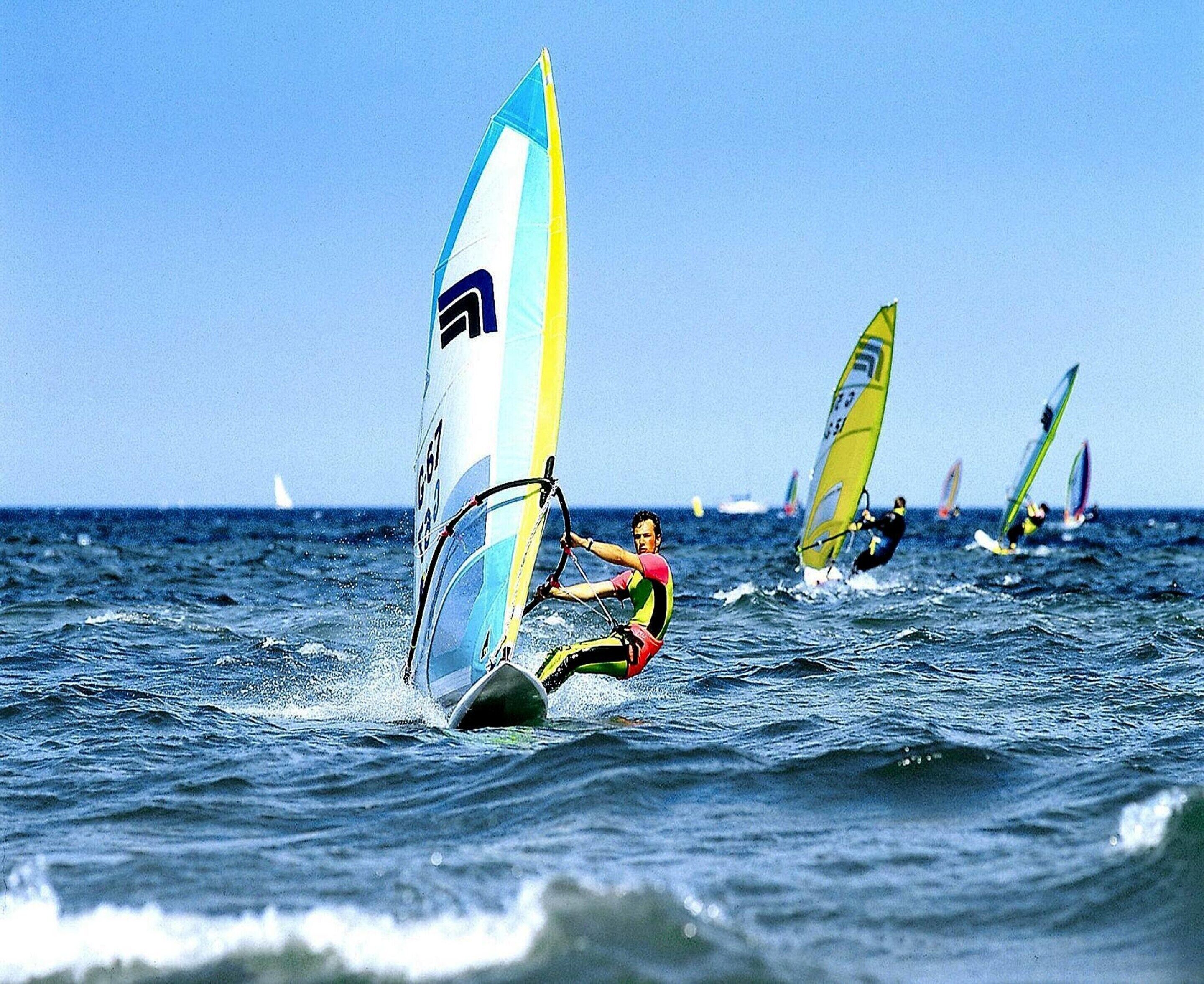Une personne pratiquant la planche à voile sur l’eau, avec plusieurs voiliers en arrière-plan.