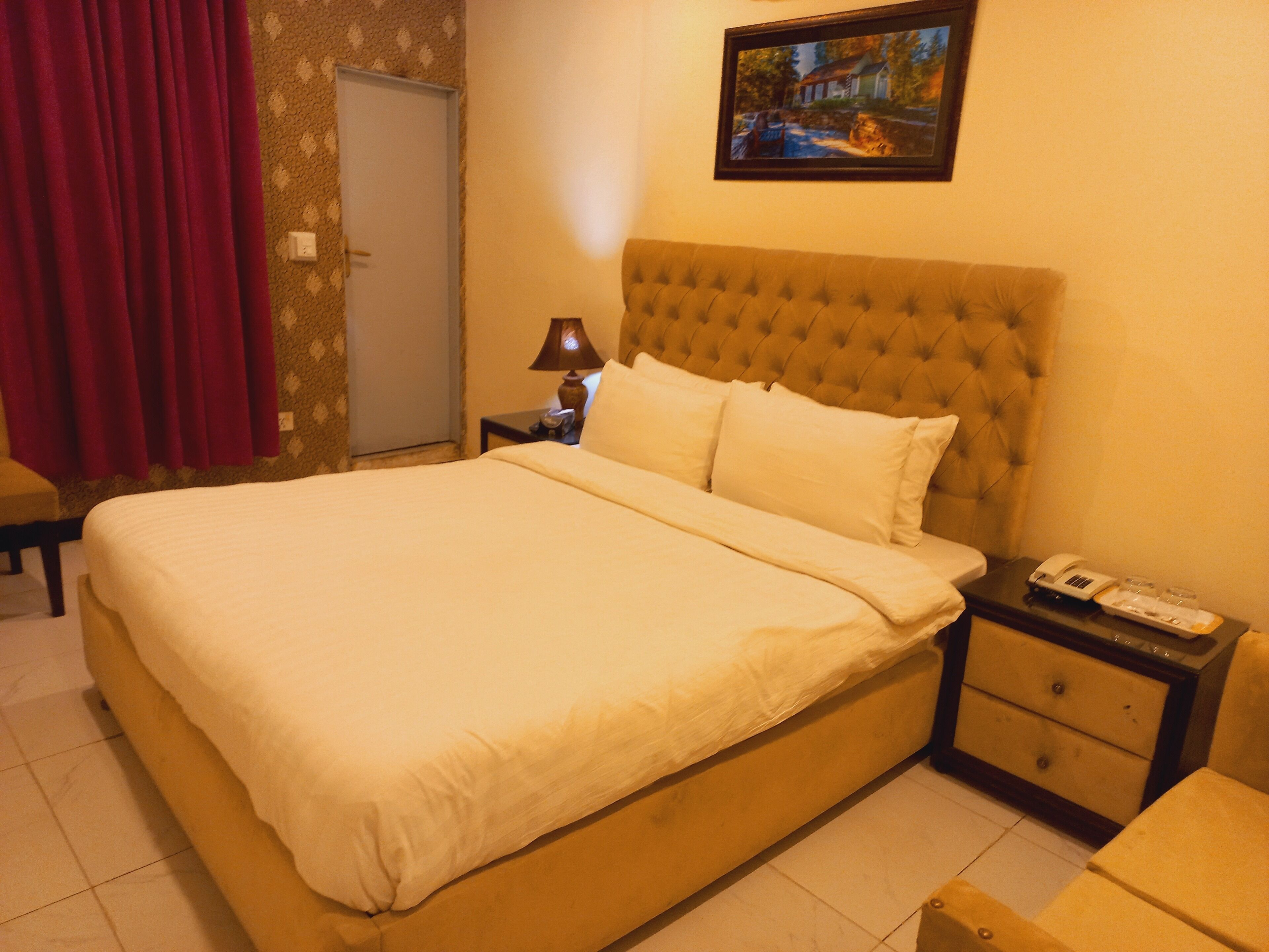 A bedroom with a large bed, a nightstand with a lamp, a telephone, and a painting on the wall.