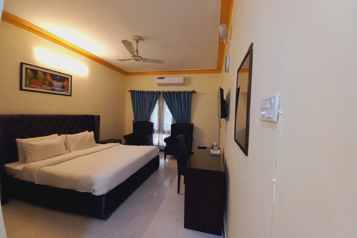 A hotel room with a bed, a desk, a chair, a ceiling fan, and a TV.