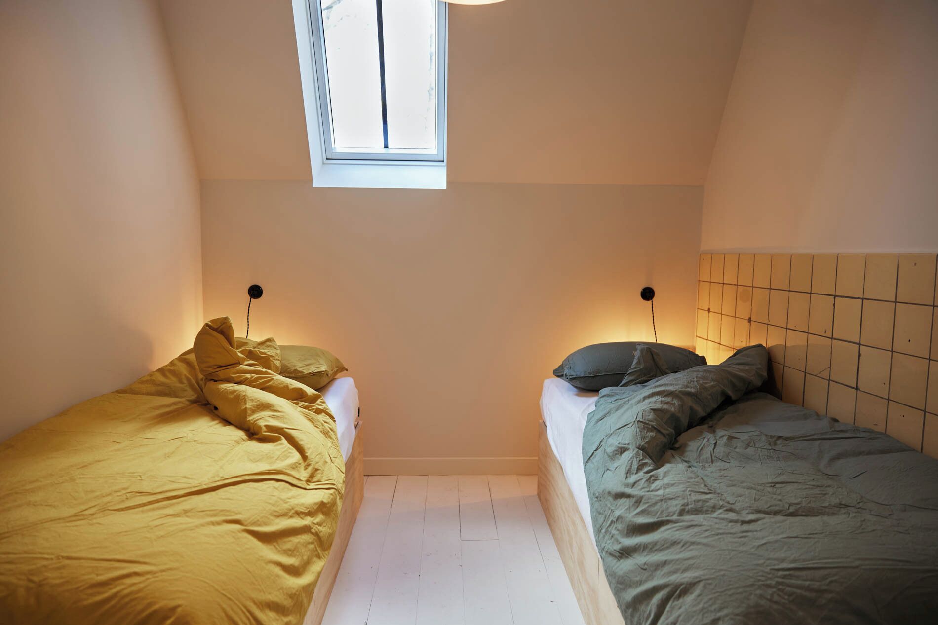 Een kleine kamer met twee eenpersoonsbedden, een raam en een betegeld hoofdeinde.