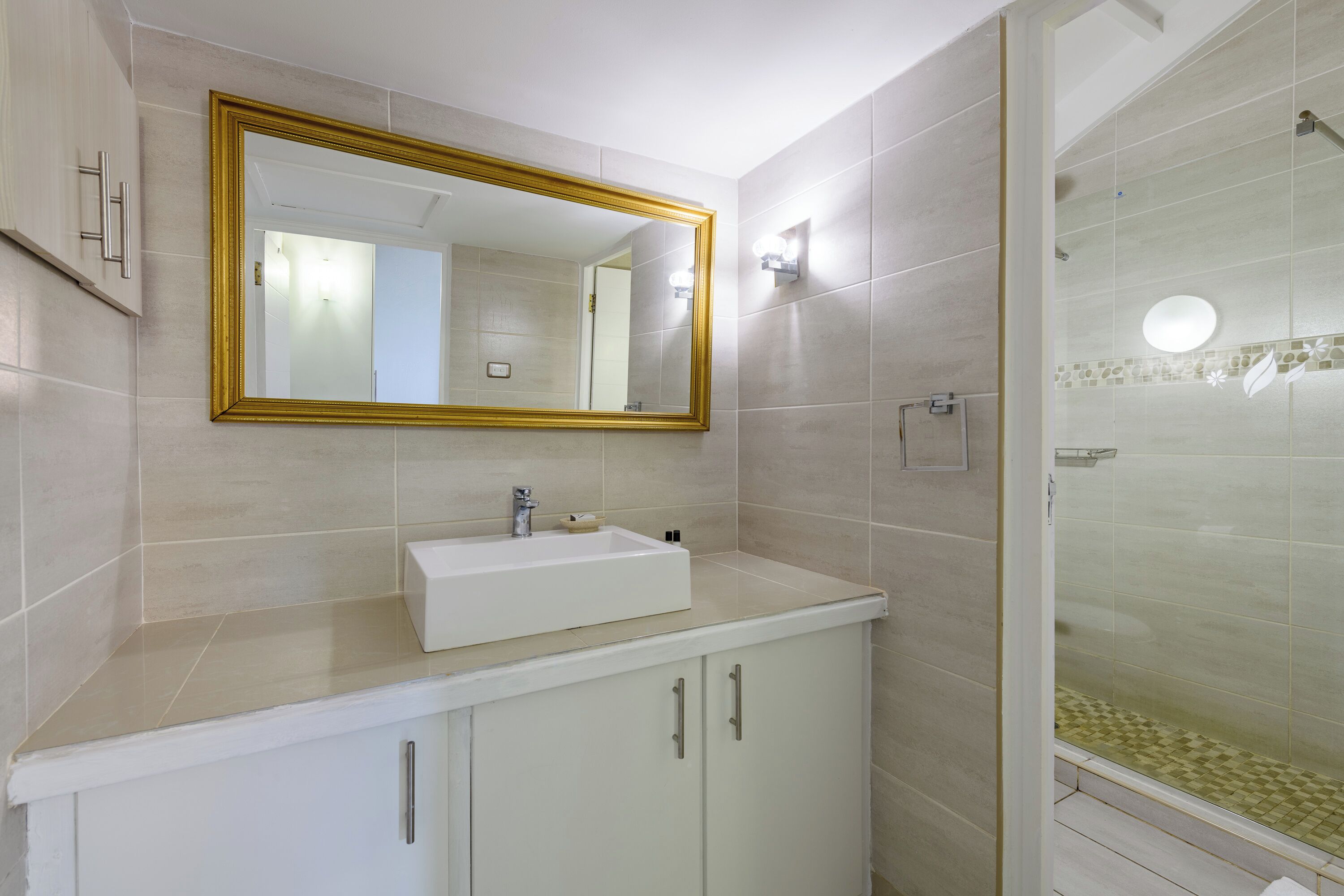 Un baño con un lavamanos blanco, un espejo con marco dorado y una cabina de ducha con paredes de vidrio.