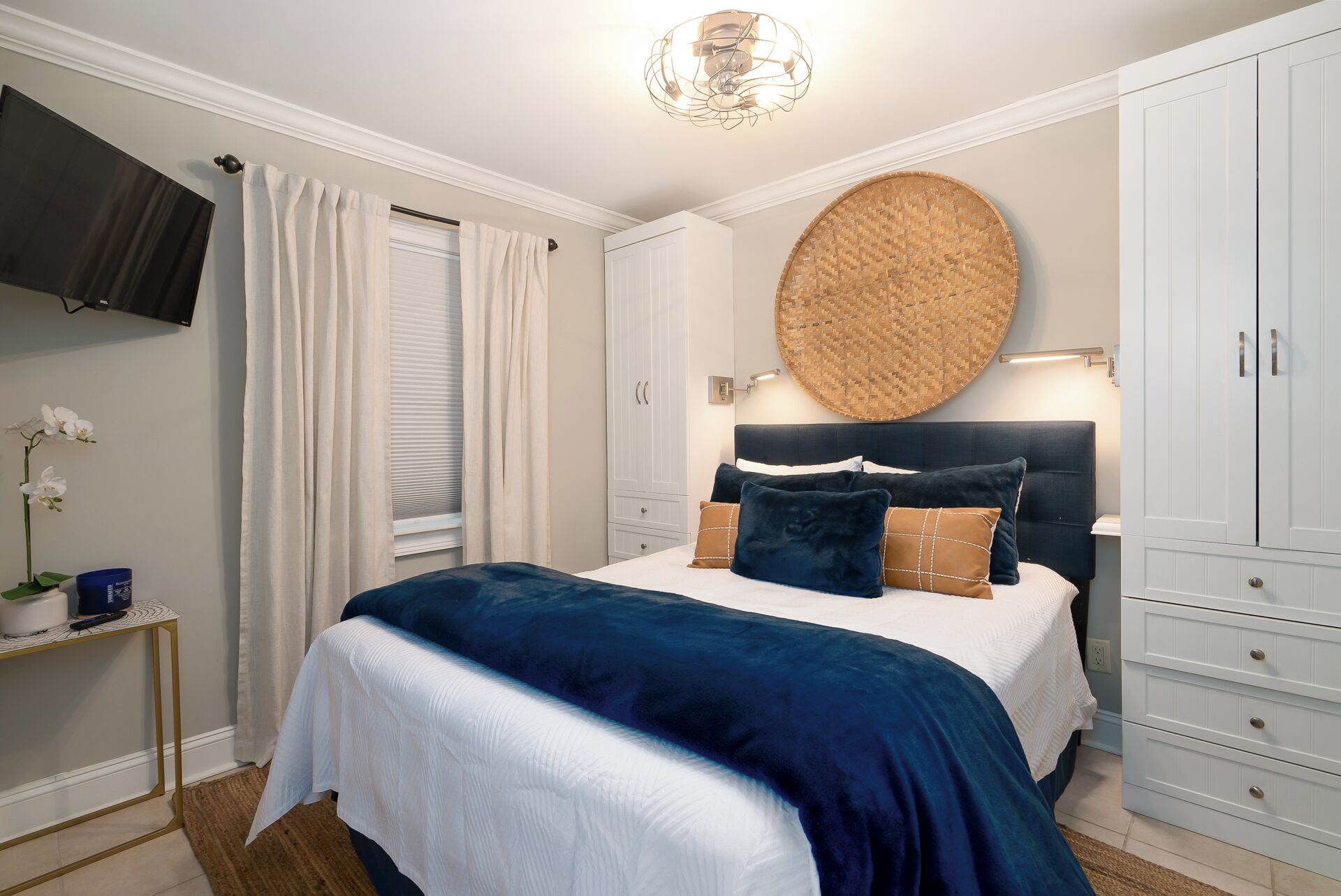 A bedroom with a bed, a round woven wall hanging, a television, and a wardrobe.
