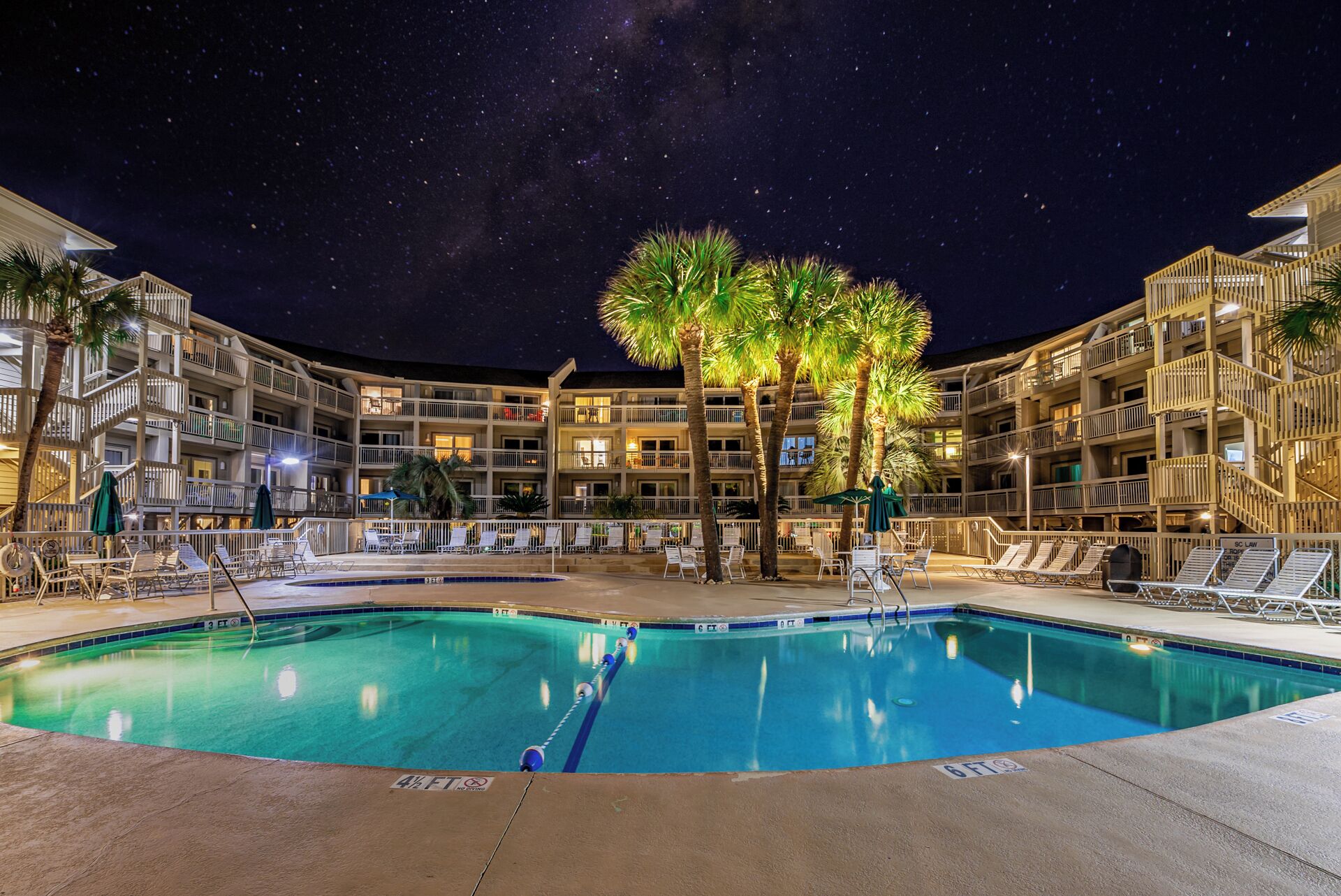 Un espace piscine nocturne avec une piscine à eau claire, des chaises longues et un immeuble à plusieurs étages doté de balcons.