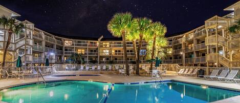 Un área de piscina nocturna con una piscina despejada, sillas de descanso y un edificio de varios pisos con balcones.