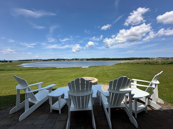 A deck with white Adirondack chairs and a stone fire pit overlooking a lake.