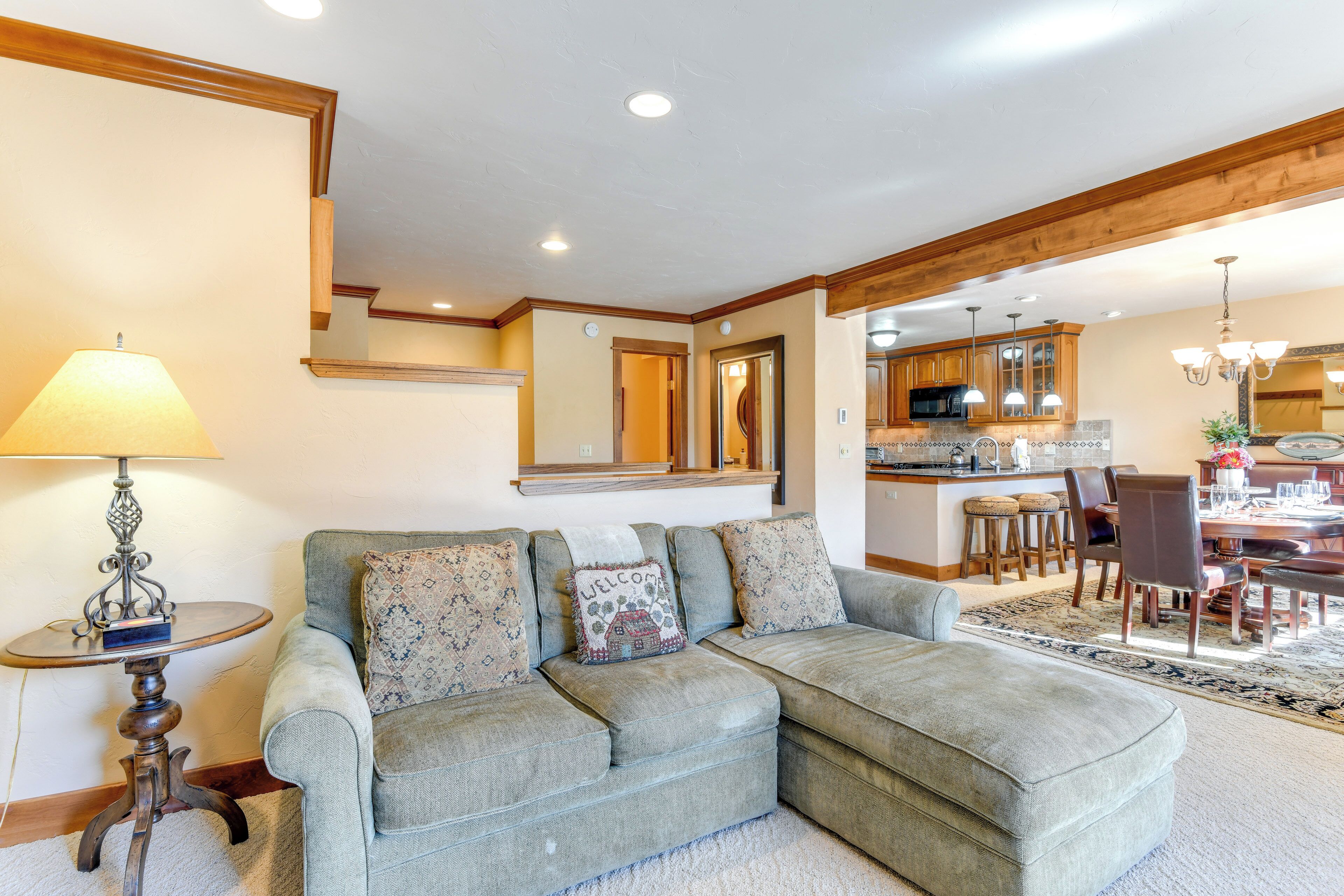 A living room with a sectional sofa, a small table with a lamp, and a kitchen in the background.