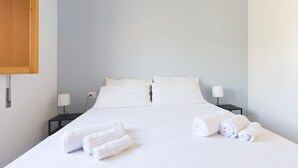 Ein ordentlich bezogenes Bett mit weißen Leinen und aufgerollten Handtüchern, eine Nachttischlampe und ein Holzschrank an der Wand.