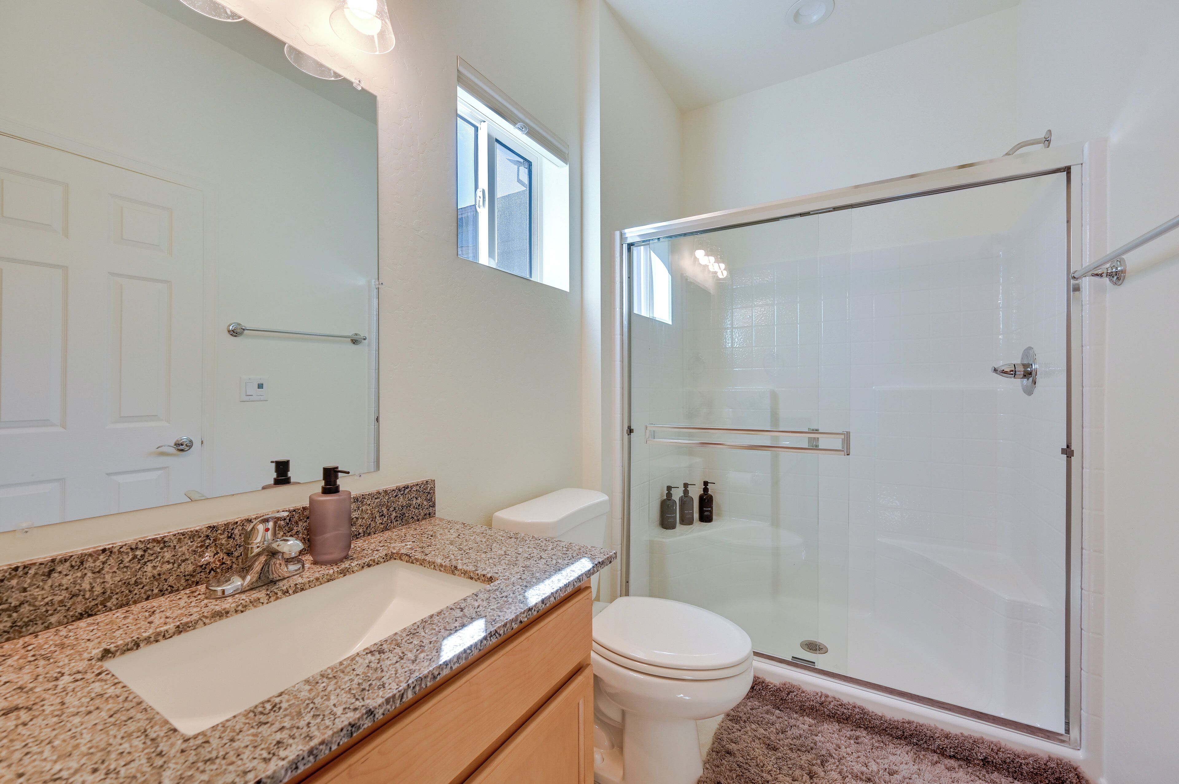 A modern bathroom with a granite countertop, a white toilet, a glass shower enclosure, and a large mirror.
