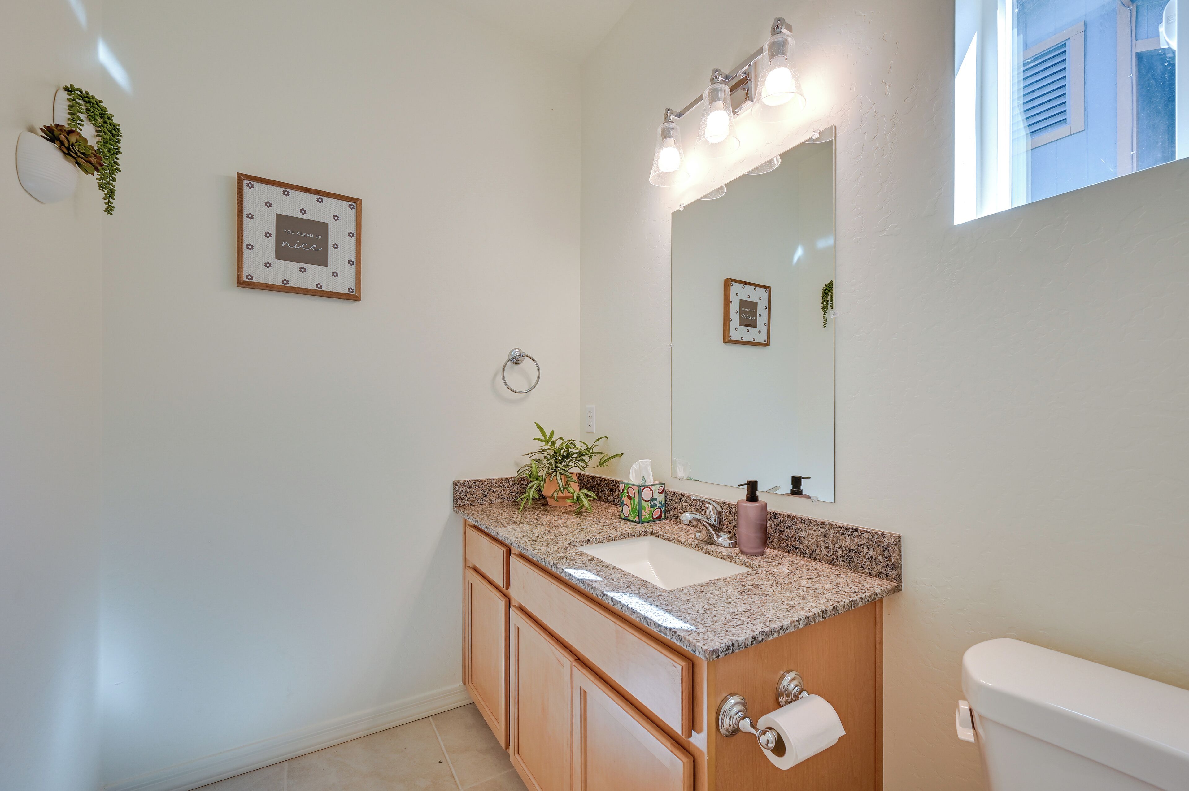 A bathroom with a sink, mirror, and toilet. There are light fixtures above the mirror and a framed picture on the wall.