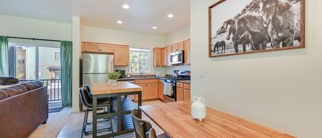 A modern kitchen with wooden cabinets, a stainless steel refrigerator, and a microwave. There is a dining table with chairs and a potted plant.