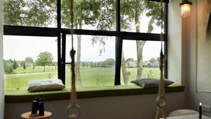 A room with a large window overlooking a green field and trees, a bedside table with a candle, and a hanging chair. - Suvelfabryk appartement 10 (Ljussens)