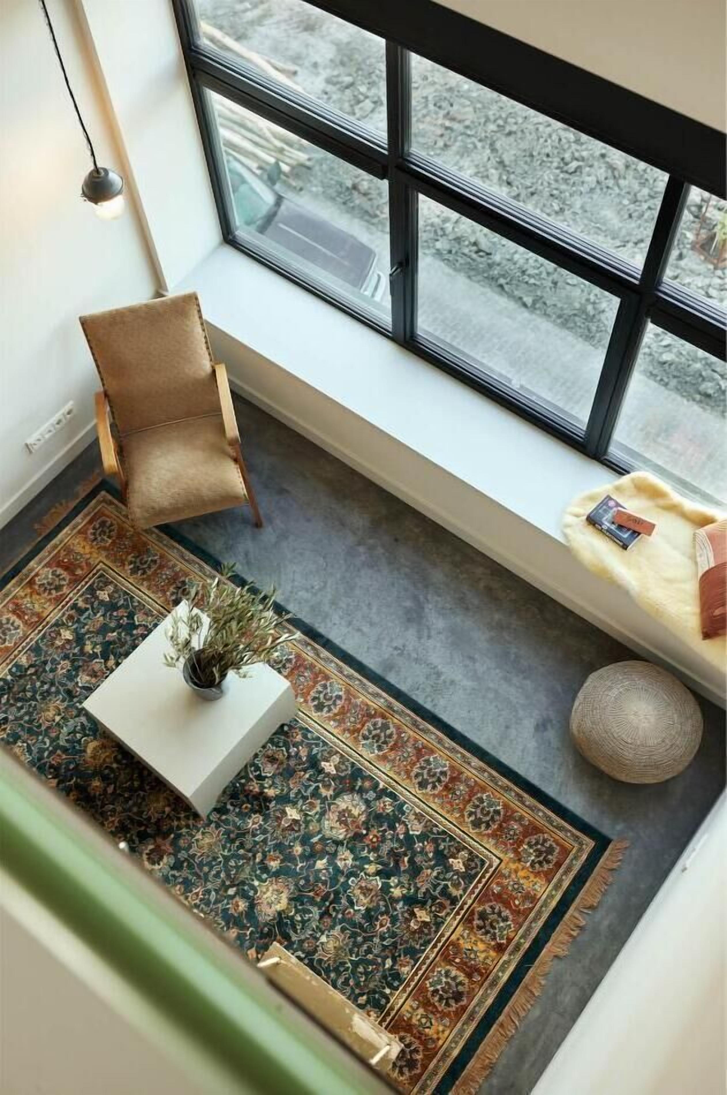 A room with a large window, a patterned rug, a white table with a plant, a beige chair, and a round decorative item on the floor.