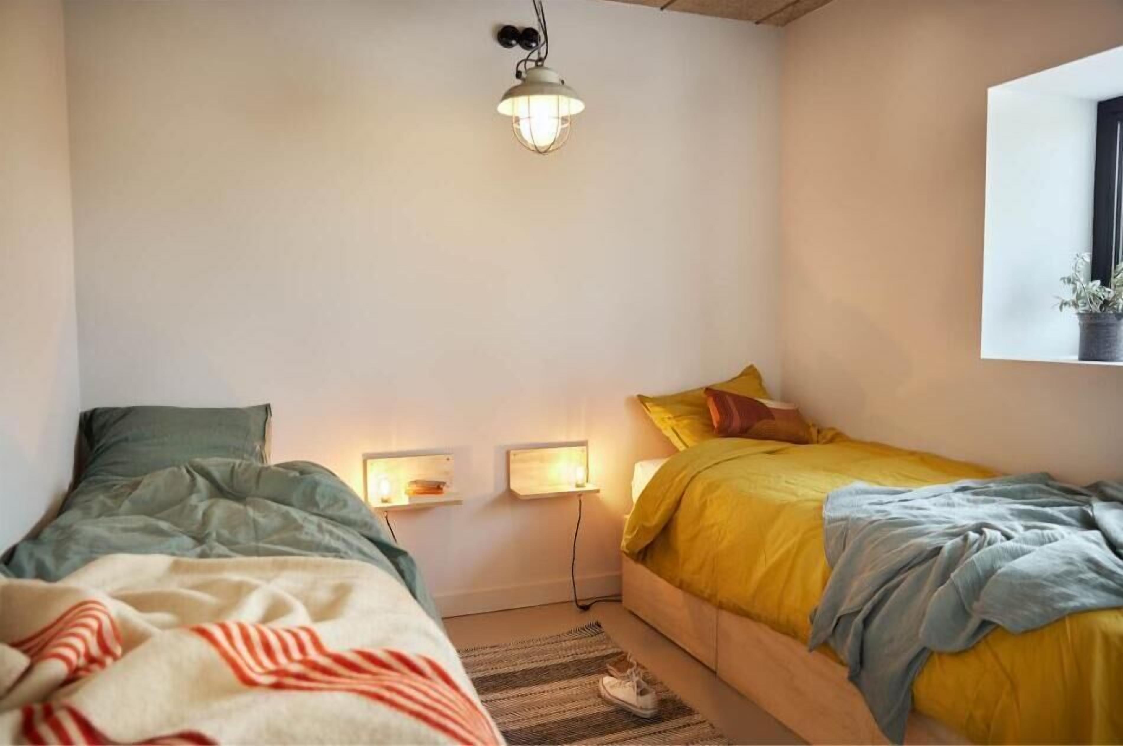 Ett sovrum med två sängar, en hängande taklampa, sänglampor och ett fönster med en växt i.