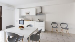 Una cocina moderna con armarios blancos, horno empotrado y estufa a gas. Hay una mesa de comedor con cuatro sillas y un bol de fruta encima.