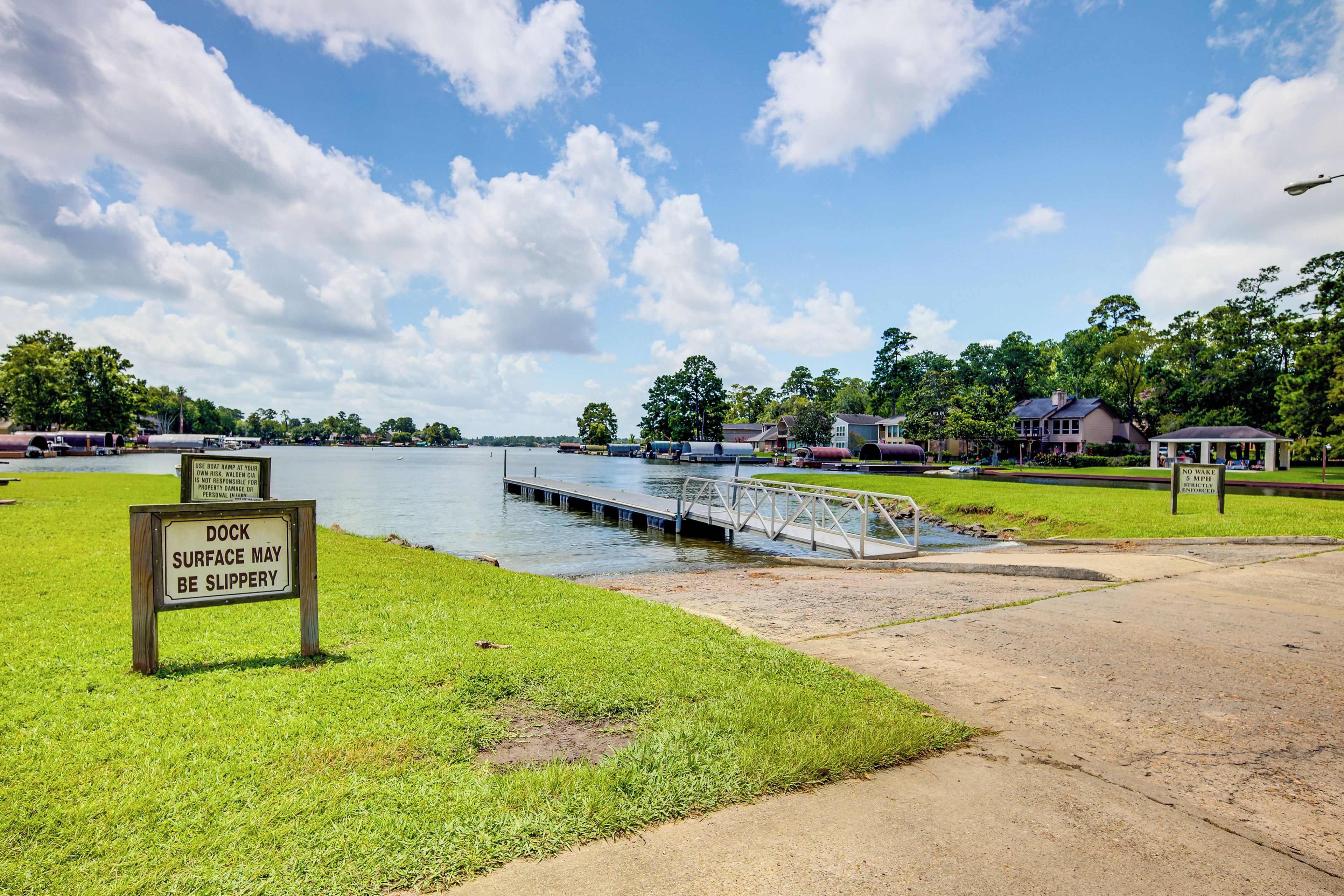 A dock with a warning sign, a grassy area, a body of water, and houses in the background.
