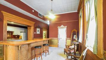 A bar area with wooden stools, a wooden counter, and a mirror. There is a door with a decorative frame and a window with curtains.