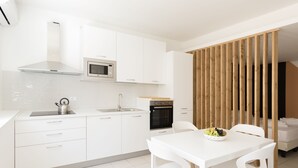 Une cuisine moderne avec des meubles blancs, un four à micro-ondes, un four traditionnel et un évier. On y trouve une table à manger avec des chaises et un saladier rempli de fruits.