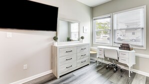 A room with a large flat-screen TV mounted on the wall, a white dresser with multiple drawers, a desk with a chair, and large windows with blinds. - 2 Blocks to Beach: Apt w/ Grill in Ocean Gate! (Ocean Gate)