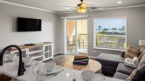 A modern living room with a flat-screen TV, a dining area with a white table set, a grey sofa, and a large window with a view of the outdoors. - New Heated Pool & Hot tub! Escape Here! (Galveston)