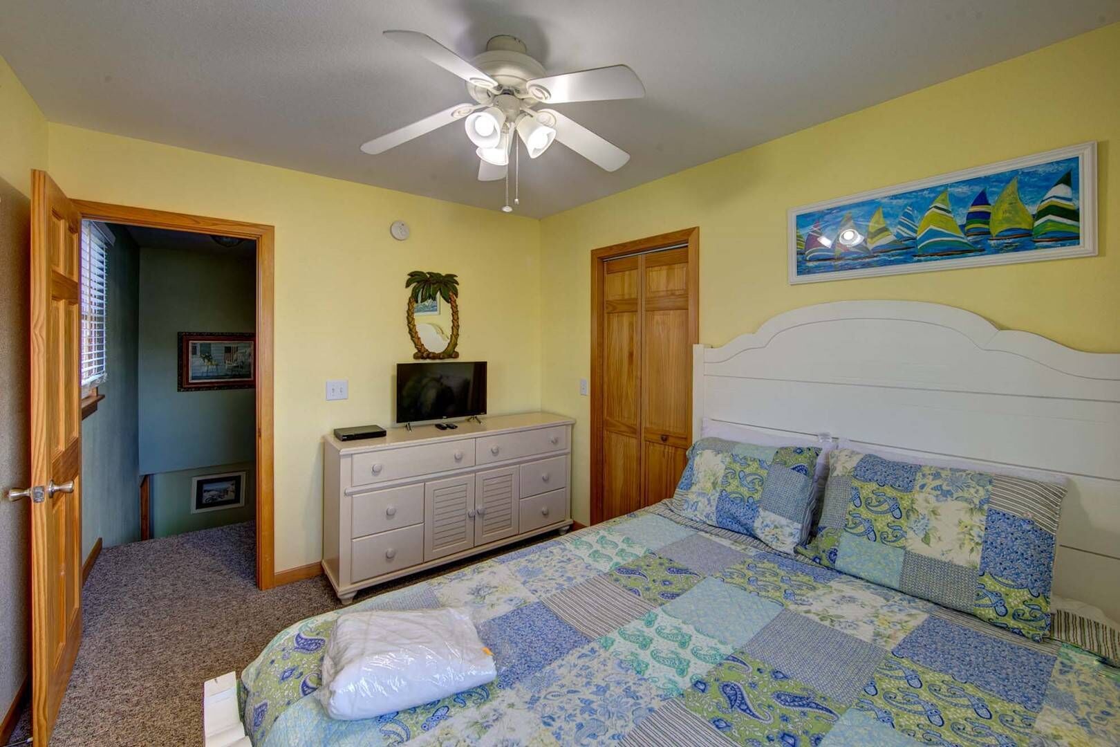 Une chambre à coucher comprenant un lit, une commode, un ventilateur de plafond, un tableau et une porte donnant sur une autre pièce.