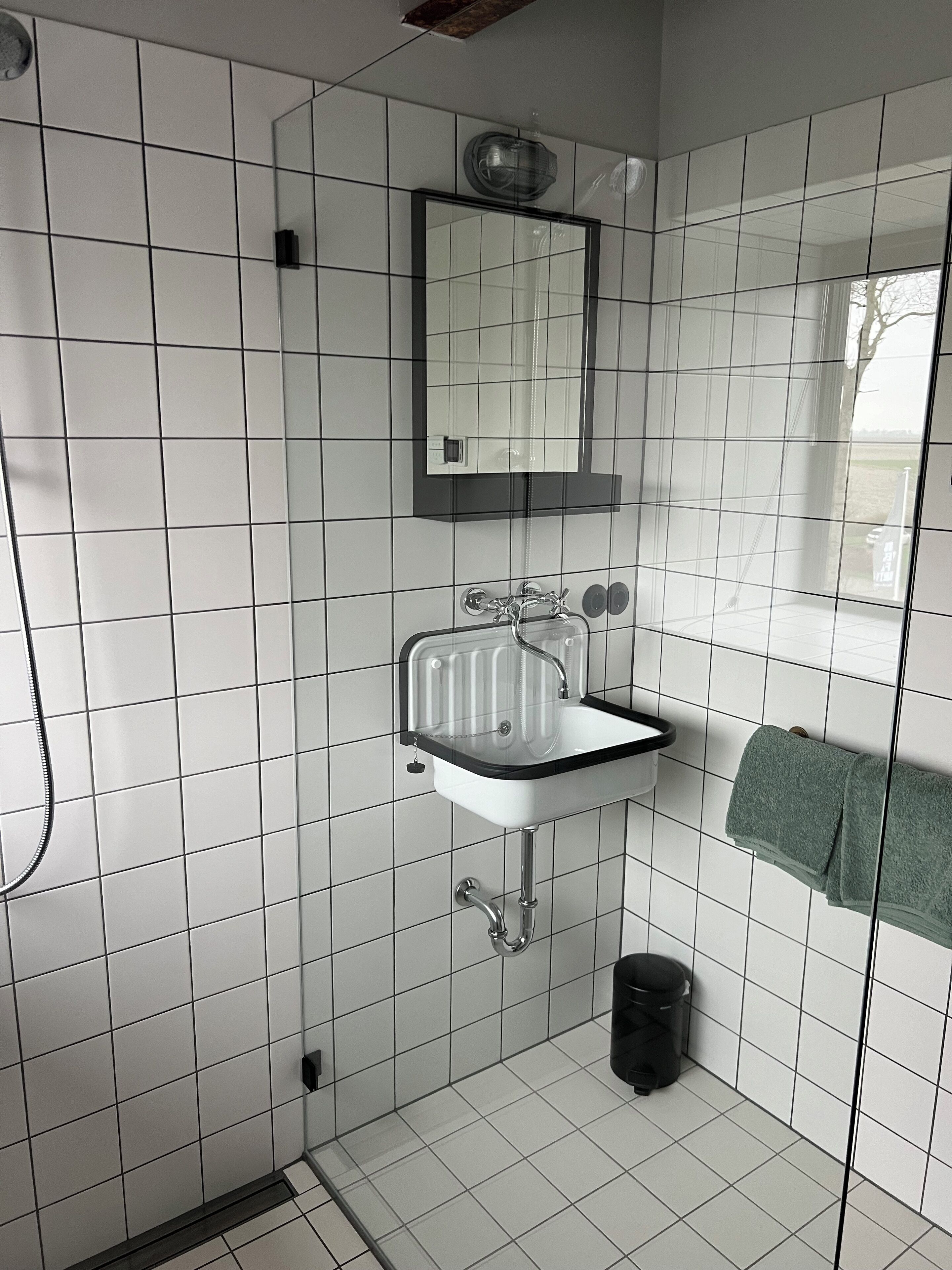 Un baño con paredes de azulejos blancos, un lavamanos blanco con encimera negra, un espejo sobre el lavamanos y un perchero con una toalla verde.