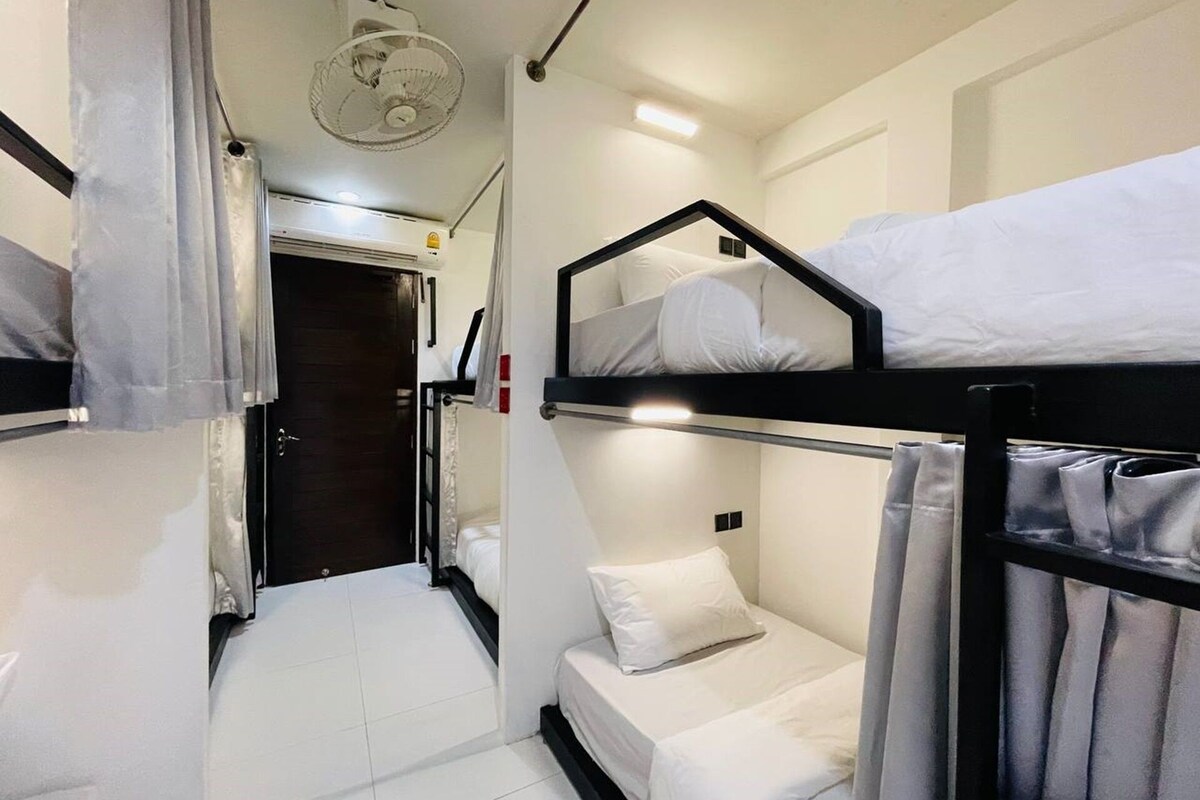 Ein kleines Hotelzimmer mit Stockbetten, einem kleinen Nachttisch, einem Ventilator und einer Tür zu einem weiteren Zimmer.