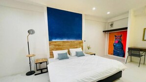 A modern bedroom with a large bed, a wooden headboard, a floor lamp, a small table, and a vibrant painting on the wall. - Samui Beach Hostel (Koh Samui)