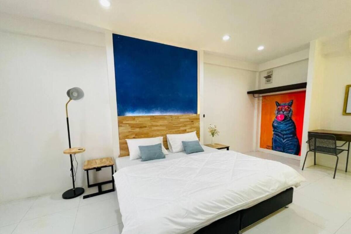 A modern bedroom with a large bed, a wooden headboard, a floor lamp, a small table, and a vibrant painting on the wall.