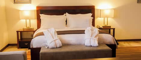 A neatly made bed with white linens and two pillows, flanked by two bedside tables with lamps, a wooden headboard, and a small ottoman.