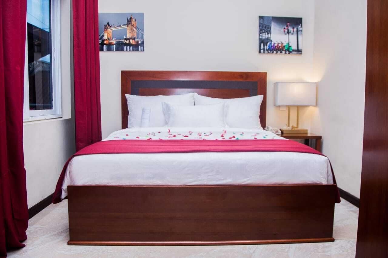 A hotel room with a wooden bed, red curtains, a nightstand with a lamp, and two wall pictures.