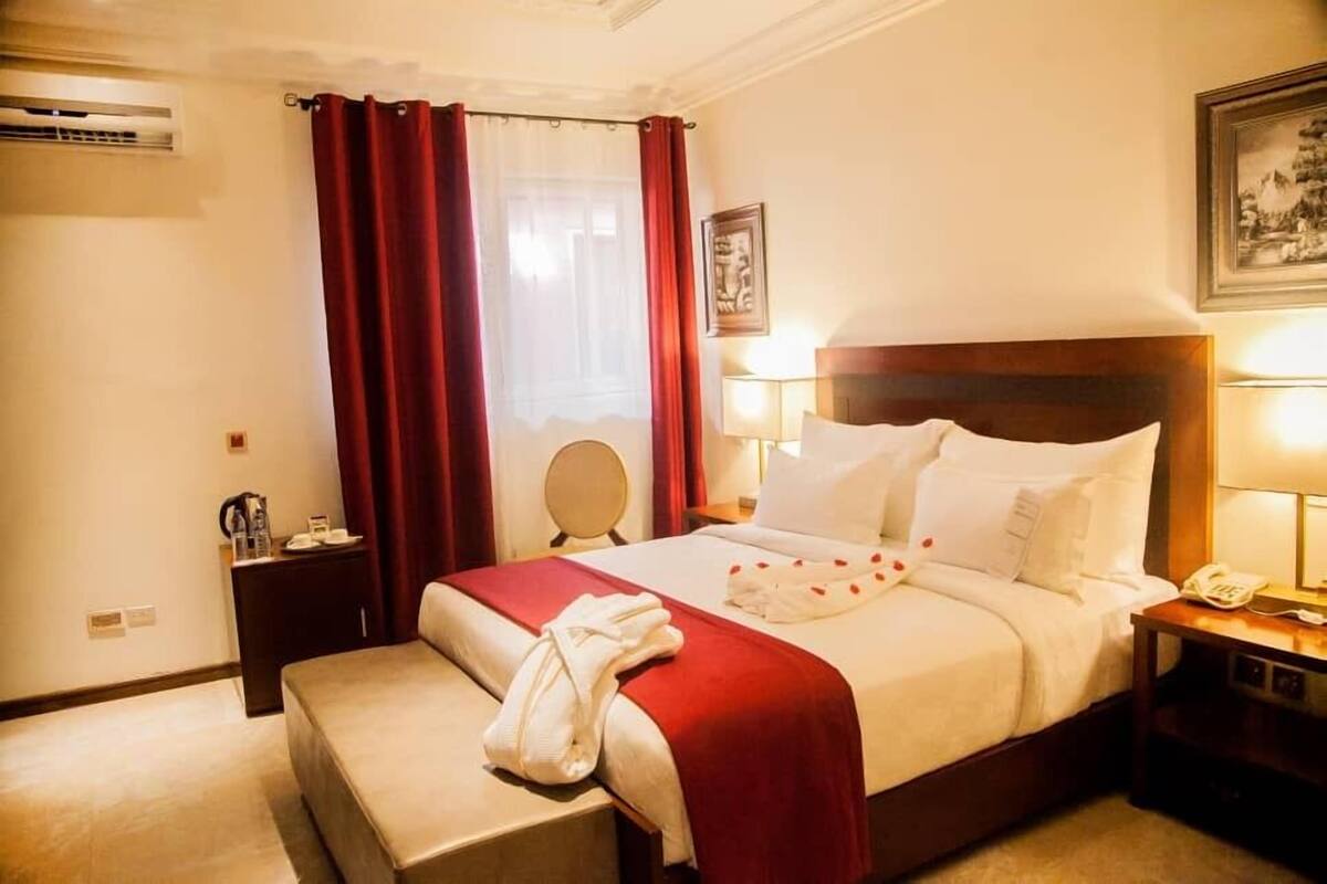 Une chambre d’hôtel comprenant un lit, une table de chevet, une commode, un miroir, une fenêtre avec des rideaux et un climatiseur fixé au mur.