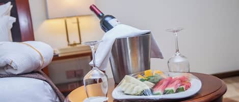 Habitación de hotel con una cama bien hecha, una mesita de noche con una botella de vino, una copa de martini y un plato de fruta.