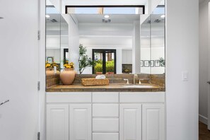 Une salle de bain moderne avec un grand miroir, des meubles blancs, un plan de travail en pierre et un vase rempli de fleurs.
